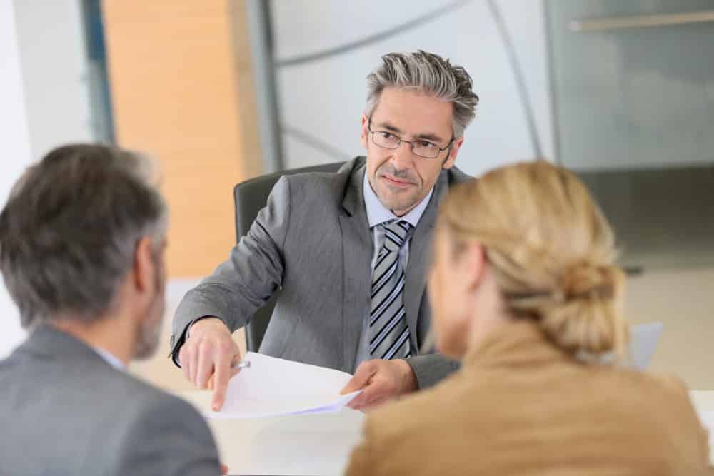 middle aged man in gray suit pointing to paper in front of couple